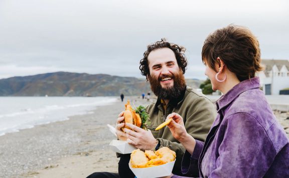 Two people enjoy a meal together on a pebble beach by the water.