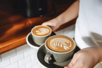 A barista at Evil Twins cafe holding two cups of coffee, both with fern designs in them.