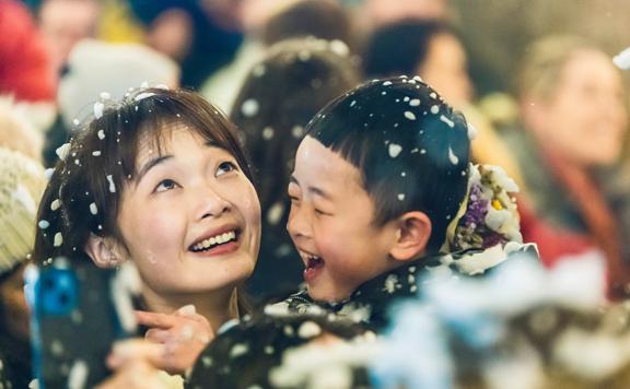 An adult holds a child, both smiling while snowflakes fall from above at the Greytown Festival of Christmas.