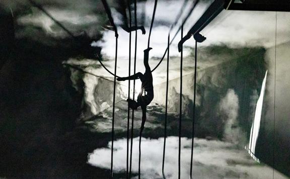 A silhouetted aerialist hangs upside down from ropes in a smoky, colourless landscape during a performance for the Aotearoa New Zealand Festival of the Arts.