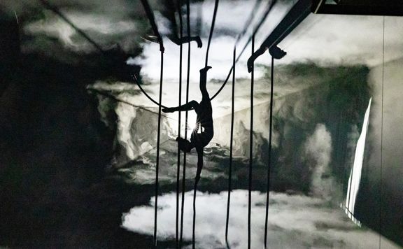 A silhouetted aerialist hangs upside down from ropes in a smoky, colourless landscape during a performance for the Aotearoa New Zealand Festival of the Arts.