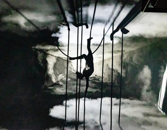 A silhouetted aerialist hangs upside down from ropes in a smoky, colourless landscape during a performance for the Aotearoa New Zealand Festival of the Arts.