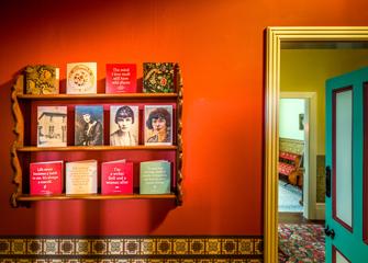 A room inside Katherine Mansfield House & Garden, in Wellington, New Zealand. There are 12 printed portraits and quotes on a wooden shelf affixed to an orange wall.