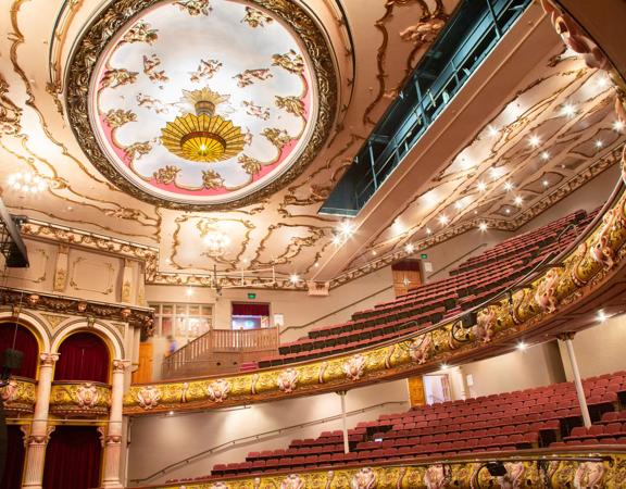 Looking up to the stands and roof of the St James Theatre, the red seats and gold fixtures can be seen, as well as the very large light on the roof.