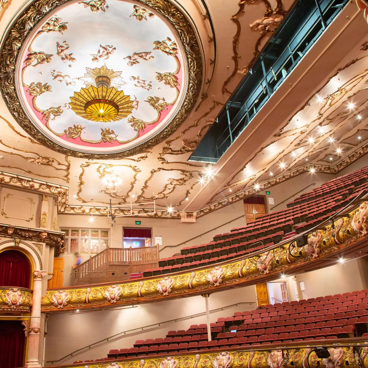 Looking up to the stands and roof of the St James Theatre, the red seats and gold fixtures can be seen, as well as the very large light on the roof.