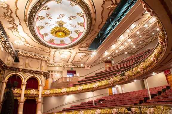 Looking up to the stands and roof of the St James Theatre, the red seats and gold fixtures can be seen, as well as the very large light on the roof.