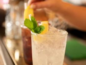 A lemonade being made at the bar of Kisa, with a lemon slice and green garnish atop. A hand in the background places a garnish on another drink.