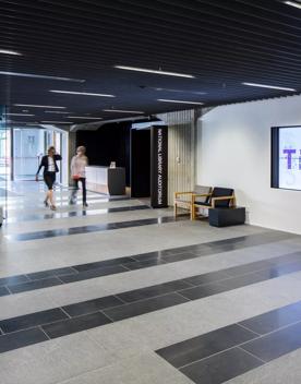 In the foyer inside Tiakiwai Conference Centre, 2 people walk past a screen and concrete tiled floors.