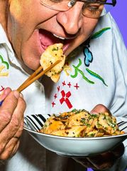 A close-up shows a person using chopsticks to lift a dumpling from a bowl of pasta or dumplings garnished with herbs.