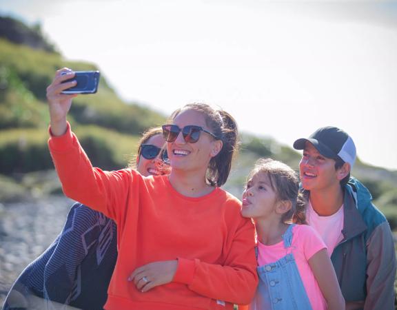 Person in a bright orange jersey holding their camera up and taking a selfie. There are three other people smiling towards the camera behind them, including a child on the right, poking their tounge out.