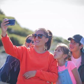 Person in a bright orange jersey holding their camera up and taking a selfie. There are three other people smiling towards the camera behind them, including a child on the right, poking their tounge out.