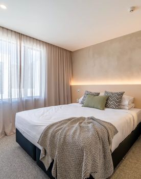 Neutral styled bedroom at NZCIS. Large bed in the centre of the room with a large window behind sheer curtains. Beige feature wall behind the bed.