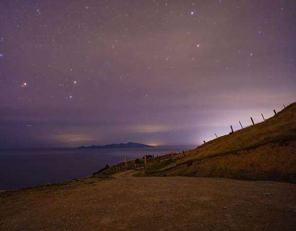 A starry night sky above a grassy hillside, the ocean and an island in the distance.