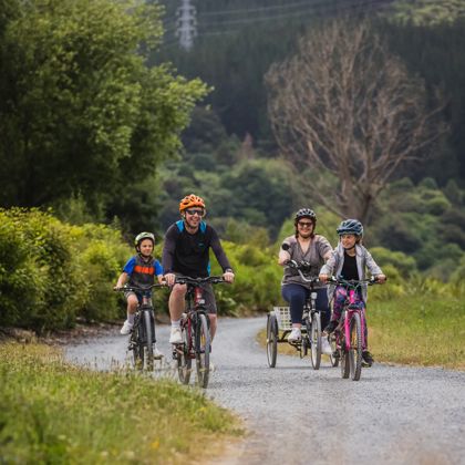 Two adults and two children riding bicycles on a gravel path, surrounded by trees.