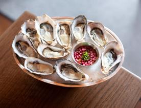 A plate of ten oysters with a small dish of mignonette sitting in  crushed ice placed on a wooden table.