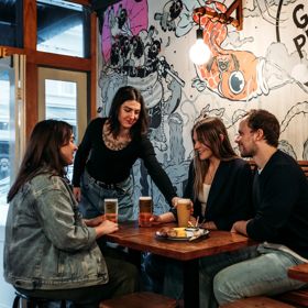A waiter hands a beer to a table of three inisde Garage Project. The wall behind them is covered in hand drawn art.
