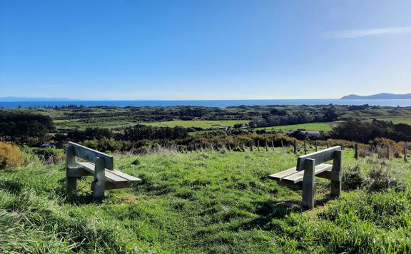 2 wooden seats atop a hill overlooking Paekākāriki, Kapiti island, and the south island in the distance.