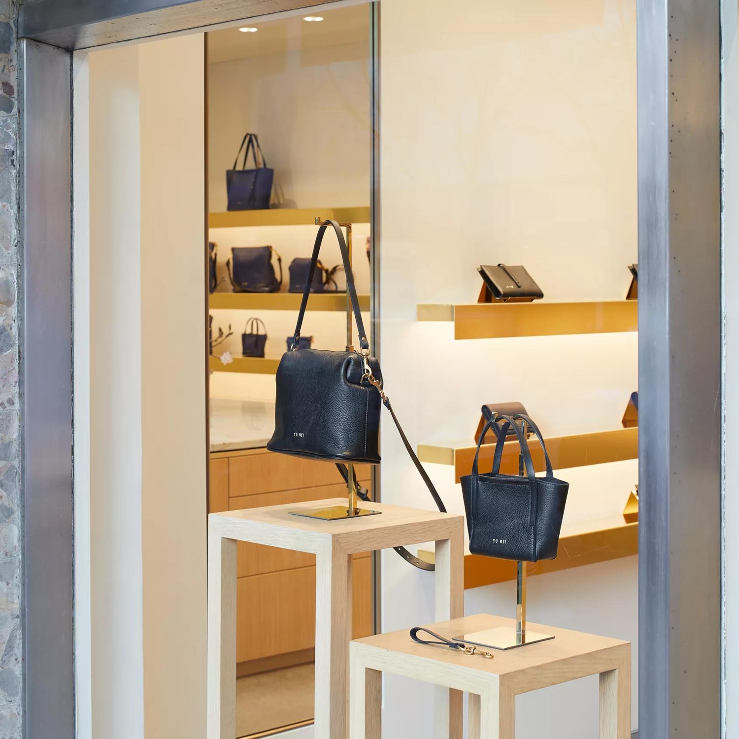 From the outside looking into Yu Mei handbag store. Yu Mei is in brass letters vertically against a terrazzo wall and two black handbags are displayed in the window.