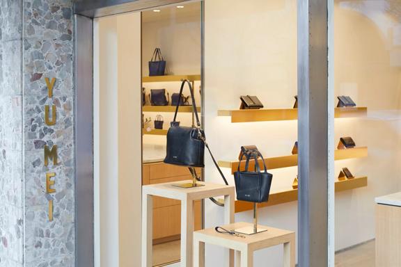 From the outside looking into Yu Mei handbag store. Yu Mei is in brass letters vertically against a terrazzo wall and two black handbags are displayed in the window.