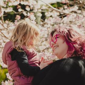 A smiling person with pink hair holds a child up, surrounded by blossoming cherry trees at Blossom Valley at Aston Norwood Gardens in Kaitoke, Upper Hutt, New Zealand. 