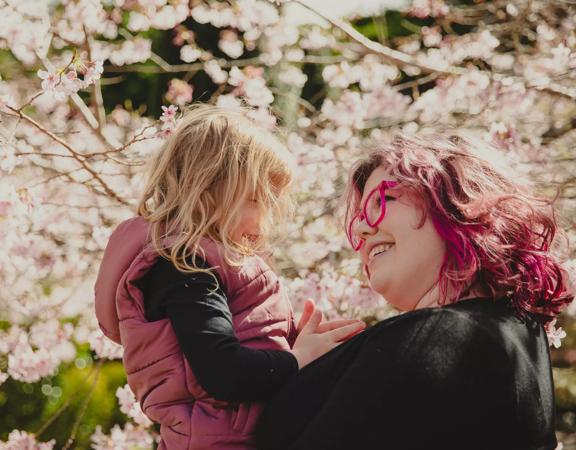 A smiling person with pink hair holds a child up, surrounded by blossoming cherry trees at Blossom Valley at Aston Norwood Gardens in Kaitoke, Upper Hutt, New Zealand. 