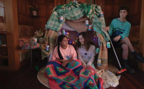 Two preteens sit in a blanket fort in the middle of a wood-panneled room looking at each other and another person sits in a chair cross-legged to the right of the fort.