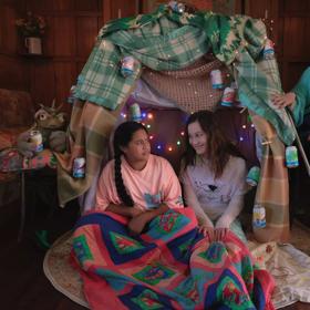 Two preteens sit in a blanket fort in the middle of a wood-panneled room looking at each other and another person sits in a chair cross-legged to the right of the fort.
