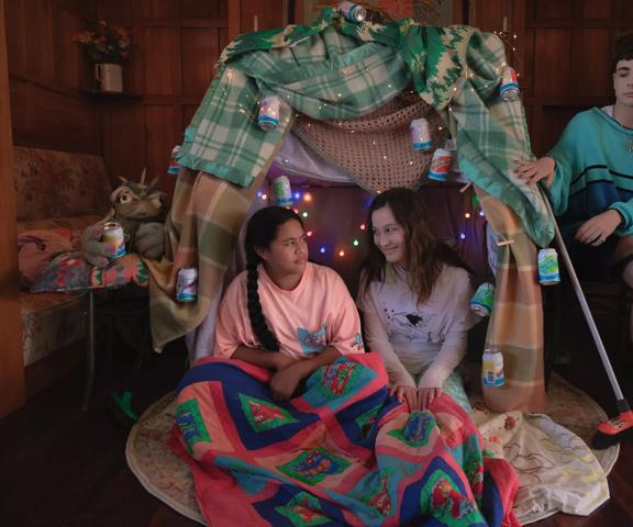 Two preteens sit in a blanket fort in the middle of a wood-panneled room looking at each other and another person sits in a chair cross-legged to the right of the fort.