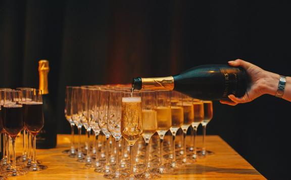 Champagne glasses lined up on a table. There is a hand holding a champagne bottle that is being poured into the glasses.