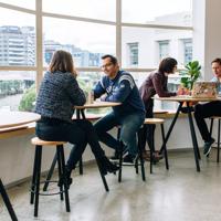 Small high tables in front of big windows at BizDojo, Wellington, with people talking over work projects.