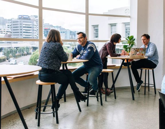 Small high tables in front of big windows at BizDojo, Wellington, with people talking over work projects.