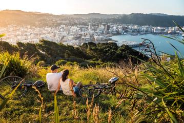 Two people sit in the grass with their bicycles lying nearby at Mount Victoria lookout in Wellington, New Zealand with a view of the city centre, the bay and rolling hills in the distance.