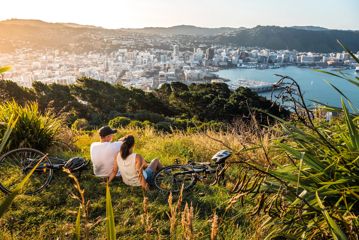 Two people sit in the grass with their bicycles lying nearby at Mount Victoria lookout in Wellington, New Zealand with a view of the city centre, the bay and rolling hills in the distance.