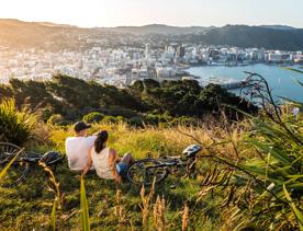 Two people sit in the grass with their bicycles lying nearby at Mount Victoria lookout in Wellington, New Zealand with a view of the city centre, the bay and rolling hills in the distance.