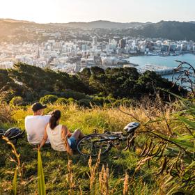 Two people sit in the grass with their bicycles lying nearby at Mount Victoria lookout in Wellington, New Zealand with a view of the city centre, the bay and rolling hills in the distance. 