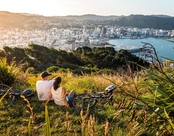 Two people sit in the grass with their bicycles lying nearby at Mount Victoria lookout in Wellington, New Zealand with a view of the city centre, the bay and rolling hills in the distance. 