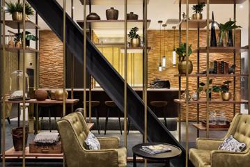 Inside Sofitel's hotel bar in Wellington, there are two olive green armchairs and a small black coffee table adorned with two magazines. The room also features decorative floating shelves, a diagonal black beam, and a bar equipped with six stools.