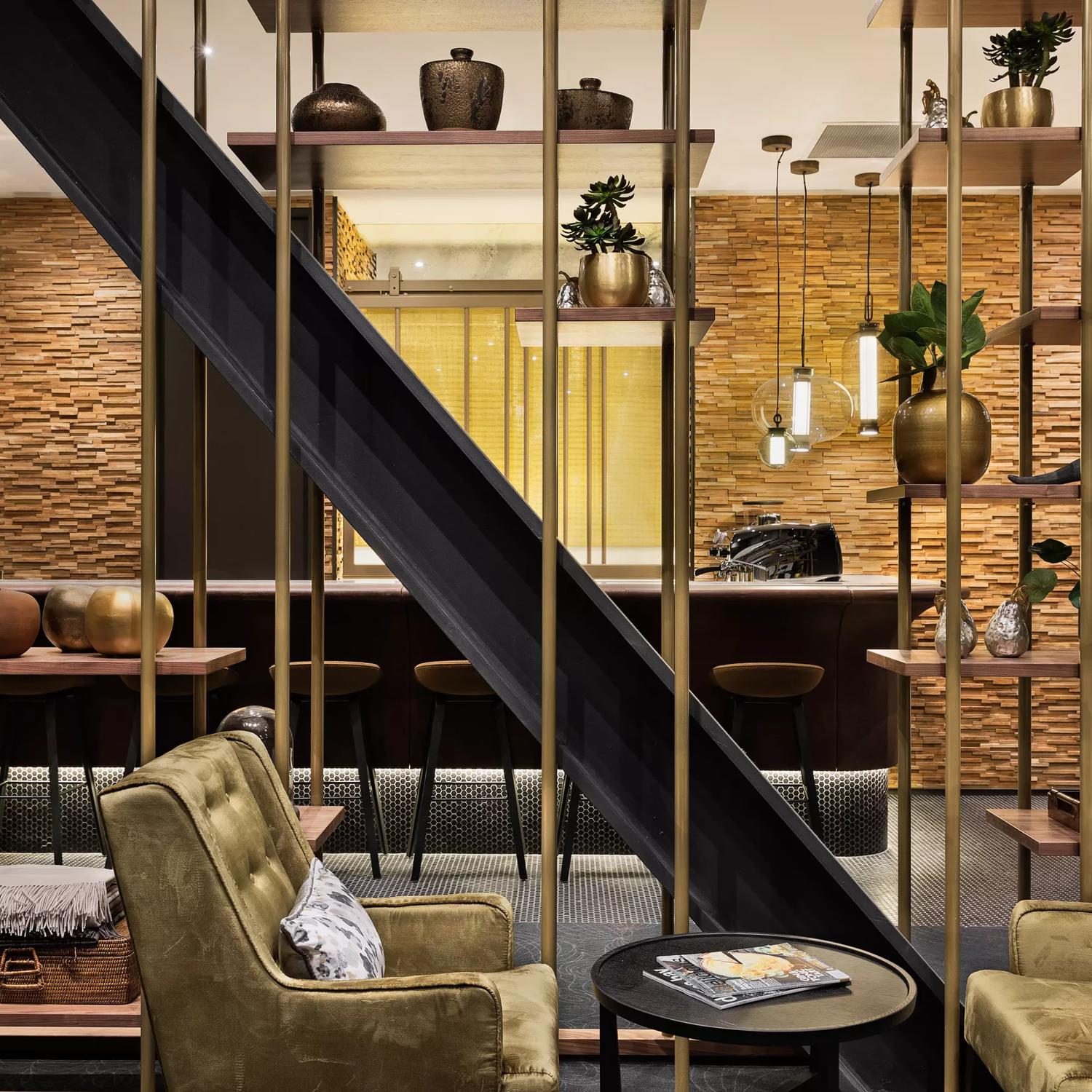 Inside Sofitel's hotel bar in Wellington, there are two olive green armchairs and a small black coffee table adorned with two magazines. The room also features decorative floating shelves, a diagonal black beam, and a bar equipped with six stools.