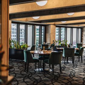 Round wooden tables and green cushioned chairs are seated in front of full-length windows at Bellamy's restaurant at the Beehive in Wellington.
