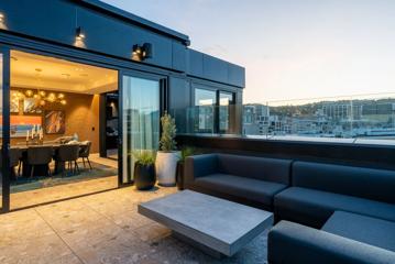 The balcony of a penthouse suite at TRYP by Windham in Wellington, New Zealand. There is a large sectional and coffee table, potted plants, and an amber-lit dining room under a dusk sky.