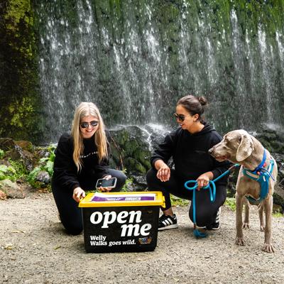 Two people and a dog are kneeling in front of a black Welly Walks box in front of Birchville Dam.