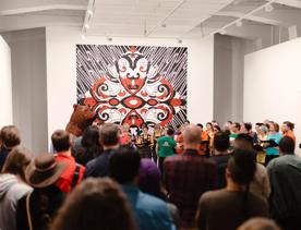A crowd of people watch a presentation in a gallery with white walls and a large-scale artwork on display. The shimmering square piece measures nearly from floor to ceiling and is a symmetrical decorative pattern using red, white and black. 