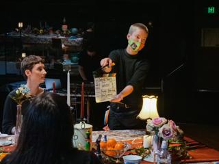 Behind the scenes on a film set where two people sit at a table and another stands holding a clap board.