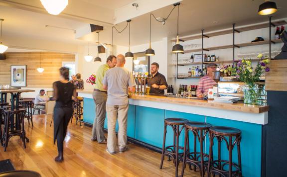 Inside the Salty Pidgin as a bartender serves patrons from behind the turquoise bar. Pendant lights hang from the ceiling.