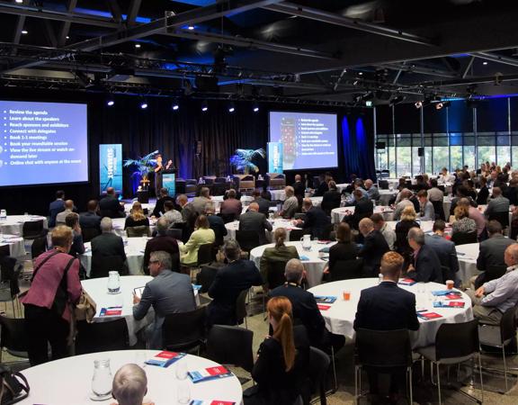 Hundreds of people seated for a conference inside the Lower Hutt Events Centre, looking at 2 large screens to the front of the room.