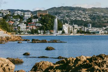 Looking towards Oriental Bay over some rocks, the fountain can be seen as well as the beach, houses, the city in the background, and Freyburg pools.