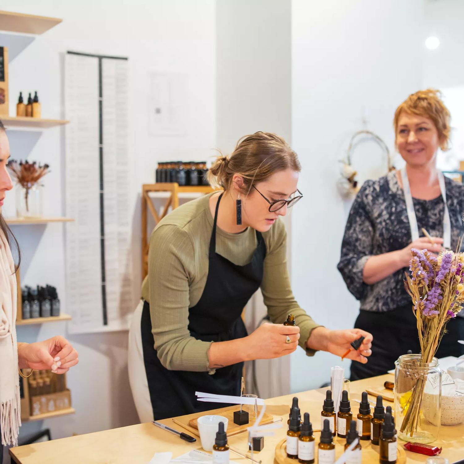 Three people stand inside the Wellington Apothecary located at 110a Cuba Street with a table with various tinctures, two mugs and a glass vase with dried flowers in front of them.