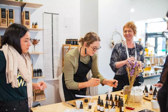 Three people stand inside the Wellington Apothecary located at 110a Cuba Street with a table with various tinctures, two mugs and a glass vase with dried flowers in front of them. 