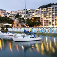 Wide shot over the wellington harbour of the Copthorne Hotel on Oriental Pde, sailboats sit on the water in front of the colourful boat sheds and the hotel behind. Photo is taken at sunset so streaks of light from the building can be seen.