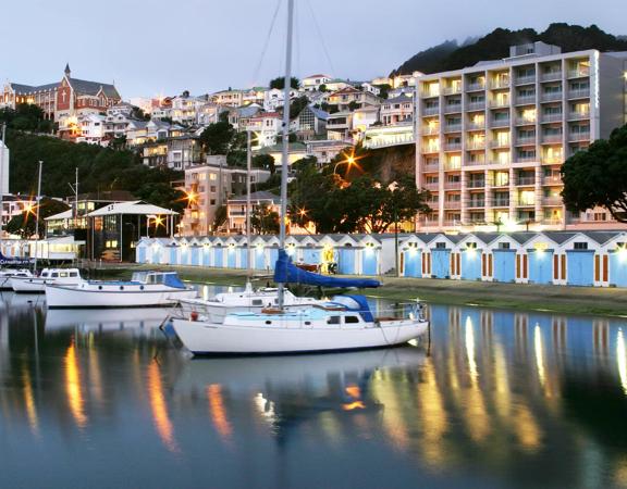 Wide shot over the wellington harbour of the Copthorne Hotel on Oriental Pde, sailboats sit on the water in front of the colourful boat sheds and the hotel behind. Photo is taken at sunset so streaks of light from the building can be seen.